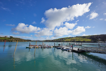 This landscape photograph captures Salcombe Harbour in Devon, England, situated in the United Kingdom. Taken during the early afternoon in the spring season, the image shows the calm waters of the harbour reflecting the clear blue sky and scattered white clouds. Several small boats are moored along floating docks, with green rolling hills of the Devon countryside visible in the background. A few people are walking along the dock and a sailboat can be seen further out, adding activity to the picturesque scene. The photograph highlights the natural beauty and tranquil atmosphere of this popular marine destination in England.